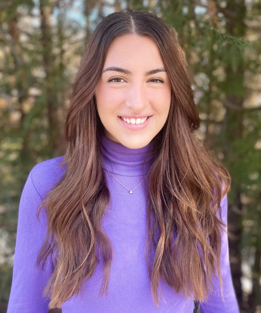 Laura is smiling, open-mouthed. She is wearing a purple turtleneck.