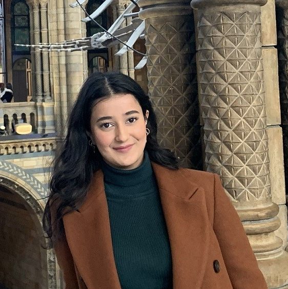 Shehnoor is smiling at the photographer. She is wearing a brown peacoat, with a green turtleneck.